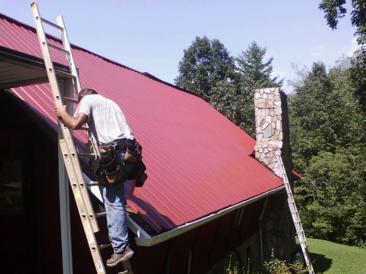 Roofer on a ladder beside a red metal roof and stone chimney — Soffit & Fascia Repair by Roofing Brothers in Saint Maries